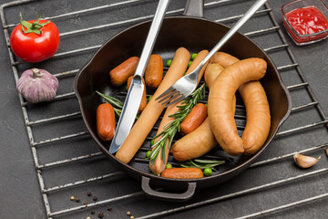 Sausages and sprigs of rosemary with knife and fork in frying pan on metal grate.