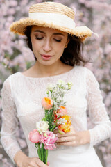 Beautiful young caucasian brunette woman with bouquet of flowers in luxury long dress among the blossoming sakura flowers. enjoying spring Early spring concept. Rest on nature vacation. straw hat