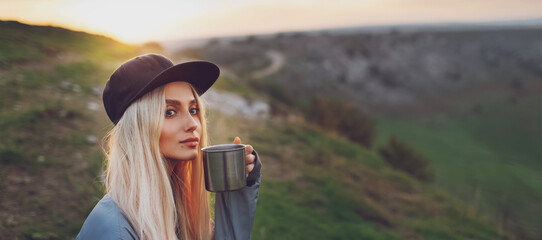 Panoramic portrait of young blonde girl holding steel cup on the peak of the hills at sunset.