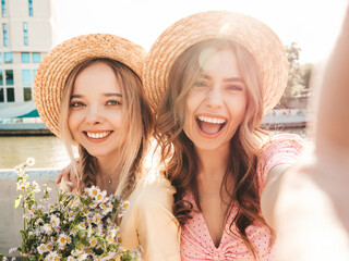 Two young beautiful smiling hipster woman in trendy summer sundress. Sexy carefree women posing on the street background in hats. Positive models at embankment at sunset. Taking selfie.Holding flowers