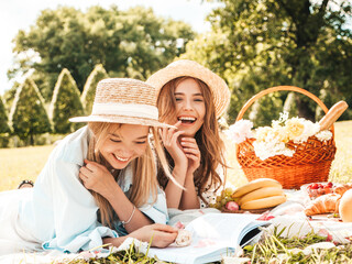 Two young beautiful smiling female in trendy summer sundress and hats.Carefree women making picnic outside.Positive models sitting on plaid on grass, reading book, eating fruits and cheese