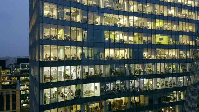 Aerial view of a skyscraper at night in winter, the drone flies around an office building from left to right office space with people working at a computer.