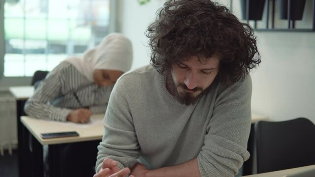 Dishonest hispanic student looking for right answer in his smartphone while writing exam test at lesson in college