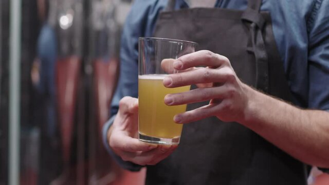 Midsection PAN shot of unrecognizable male brewery worker in apron holding glass of freshly made beer standing at beer production factory