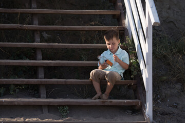 the boy is sitting on the wooden steps, wearing shorts and a T-shirt, and looking at a glass bottle