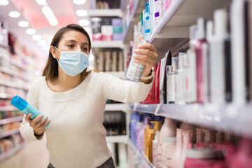 Young woman in mask choosing hair care products at shop