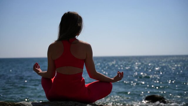 A young woman in red sportswear relaxes while practicing yoga on the beach by the calm sea, close-up of hands, gyan mudra and lotus pose.