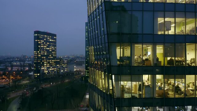 Drone shot at the office windows of a skyscraper at night, the camera moves from bottom to top panning the office windows and partially the city of Warsaw in Poland.
