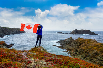 Adventurous woman holding a Canadian Flag on a Rocky Atlantic Ocean Coast during a cloudy and blue sky day. Taken in Sleepy Cove, Crow Head, Twillingate, Newfoundland, Canada. © edb3_16