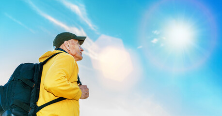Old hiker in yellow hoodie and backpack stands in mountain against blue sky