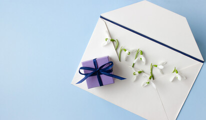 Beautiful snowdrops in white envelope and purple gift box on light blue background, closeup, space for text, top view.