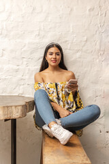Young hispanic woman with long black hair, casually dressed, looking straight at the camera while holds her smart phone and sitting at a rustic wooden table with a white brick wall in the background