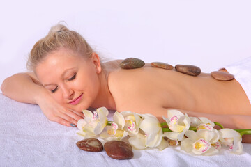 Obraz na płótnie Canvas A young girl relaxes in a spa by lying on her stomach next to orchid flowers and heated stones