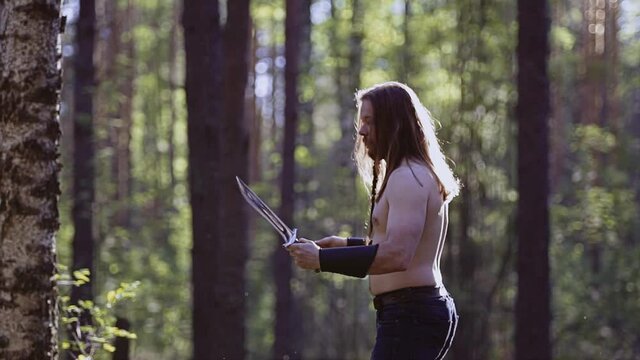 The long-haired guy throws a large knife from hand to hand, practicing techniques. Shooting in the forest