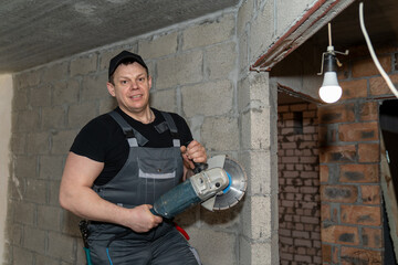 an electrician in overalls with a grinder in his hands stands against a brick wall