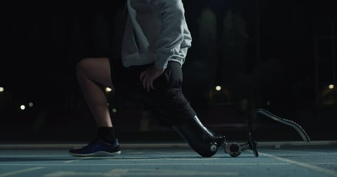 Cinematic close up shot of disable man with legs prosthesis is warming up before run with dedication on car track at night. Concept of handicapped people active lifestyle, determination, motivation.