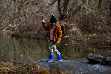 Obraz premium The girl in rubber boots in a brown jacket crosses the river ford, in early spring the trees around without leaves. Fun childhood