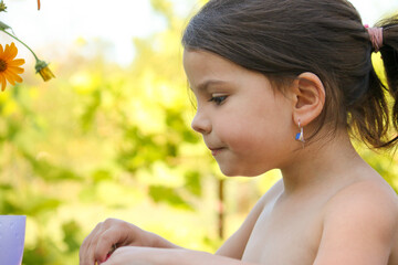 Fototapeta premium close-up portrait of a serious little girl with a ponytail on summer nature against a background of blurry green leaves