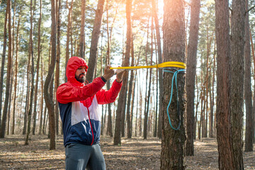 Athletic bearded man pulls the resistance band in a morning forest.
