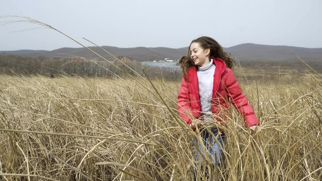 Close-up. A girl in a red jacket rejoices and jumps in the high dry grass in the field. The child frolics in the field among the tall weeds of ears.