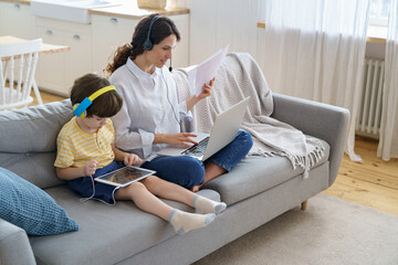 Freelancer mother sitting on couch at home office during lockdown, remote work on laptop. Child...