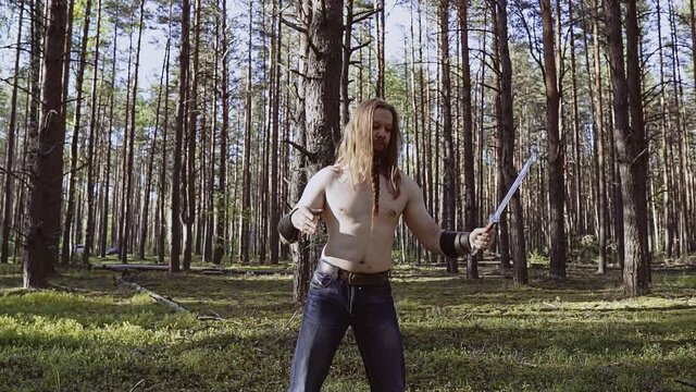 A man with a bare torso trains with a knife in his hands in the forest. Shooting a workout in motion