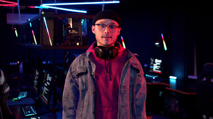 A cute young gamer in glasses and a knitted hat smiles against the background of the reception desk in the computer club