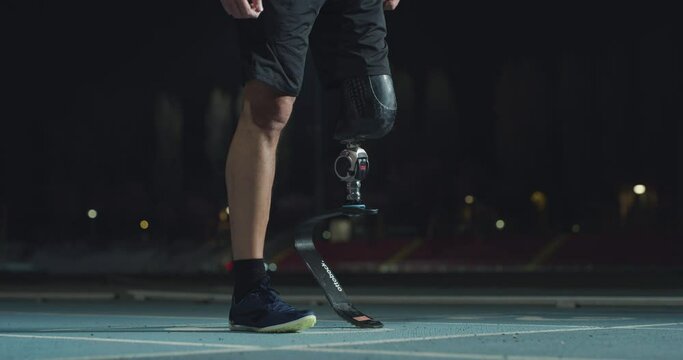 Cinematic close up shot of disable man with legs prosthesis is warming up before run with dedication on car track at night. Concept of handicapped people active lifestyle, determination, motivation.