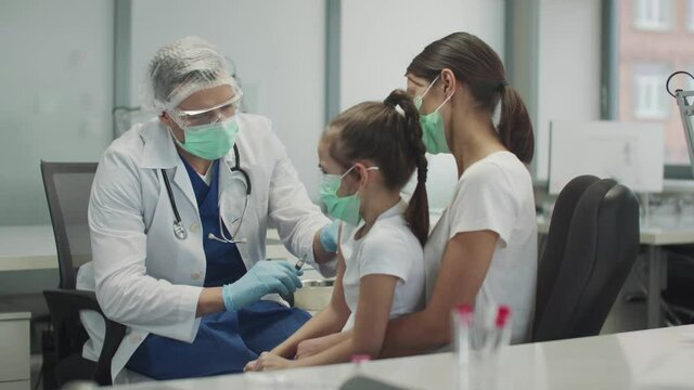 A kind doctor in protective glasses and a mask has given the child a girl a shot in the arm and nods approvingly to her, slowmotion.