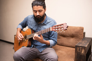 Man dressed in casual clothes playing guitar, sitting on an armchair in a room