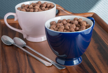 Flakes, chocolate balls in large pink and blue porcelain mugs and long small spoons on a wooden breakfast tray.