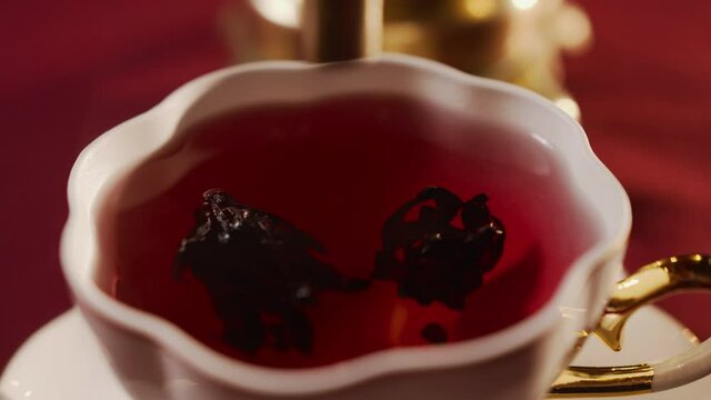 In white ceramic cup, tea is brewed from dried perianth flowers of roselle Hibiscus sabdariffa. Sour-tasting drink of bright red color. Close-up of stream of water from spout of kettle or samovar