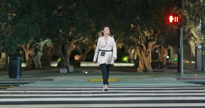 Independent woman in stylish designer jacket crossing road in night city. Confident blonde model in fashionable outfit and modern shoes walks catwalk with urban park on background. Night scene footage