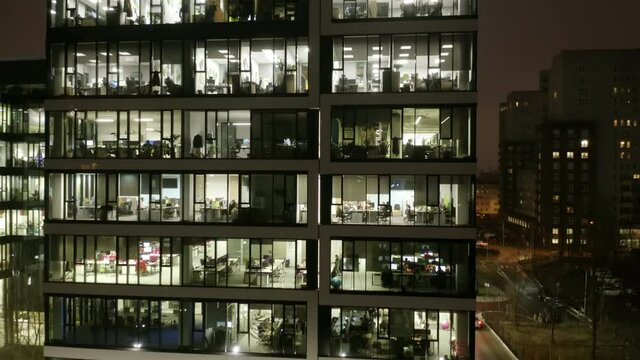 Drone shot at the windows of offices in a skyscraper at night in winter. The city of Warsaw, Poland. A cinematic view from a drone of offices in a skyscraper with people working at computers at night 