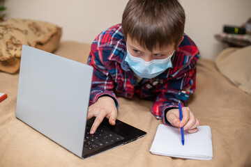 Naklejka premium boy in protective medical mask lies near laptop and writes with left hand in notebook. child does lessons watching online broadcast lessons on Internet. e-learning, remote studying internet concept