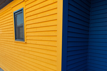 A bright yellow wooden building made of clapboard siding with royal blue colour trim. There's a single blue closed double hung window to the right. The building is on an angle with a blue sky on top. 