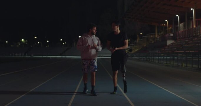 Cinematic shot of young happy friend coach is satisfied with results of disabled male runner with legs prosthesis on race track at night.Concept:handicapped people active lifestyle,friendship, victory