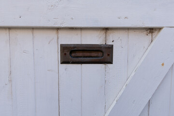 A vintage exterior wooden white door with wood panels. There's a small brass post letter or mail slot at the top and center of the wood weather shutter door. The material is worn with specs of rust.