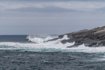 Fototapeta premium A rugged coastline on a sunny day with blue sky and clouds. The ocean is raging and is hitting the side of the craggy cliff. There's the land in the background and the teal ocean in the foreground.