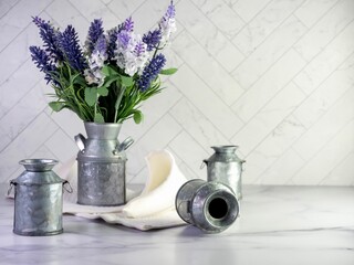 Rustic metal milk can filled with artificial purple lavender and lilacs for a simple spring country home decor with still life photography and a marble counter with a herringbone tile background.
