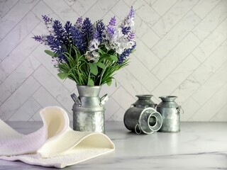 Rustic metal milk can filled with artificial purple lavender and lilacs for a simple spring country home decor with still life photography and a marble counter with a herringbone tile background.