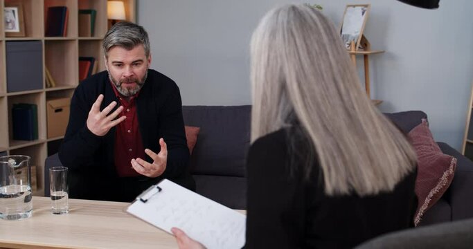 Crop view of mature man sitting on couch and having therapy session with female person. Male bearded person talking emotionally and gesturing while woman psychologist making notes.