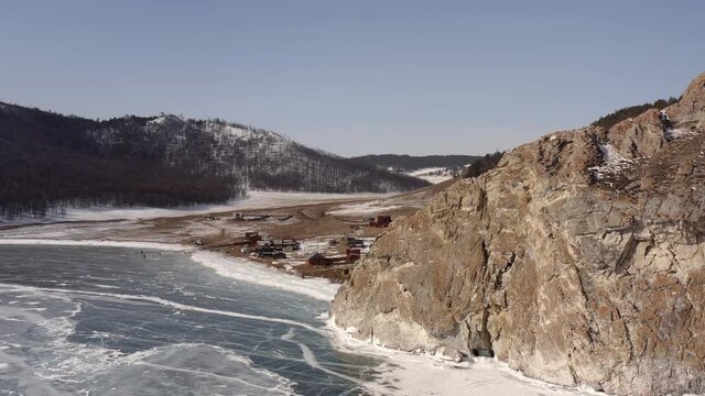 Aerial view, the drone lands near the ice, over the village and the rocky coastline. Uzury village with a coastline of rocks and hills covered with snow. Frozen Lake Baikal