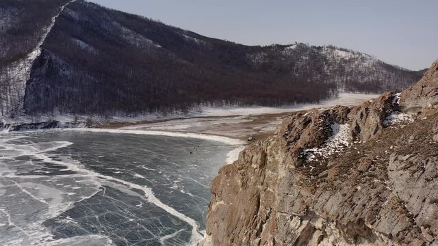 Aerial view, the drone takes off near the ice, over the rocky coastline with an opening view of the village. Uzury village with a coastline of rocks and hills covered with snow. Frozen Lake Baikal