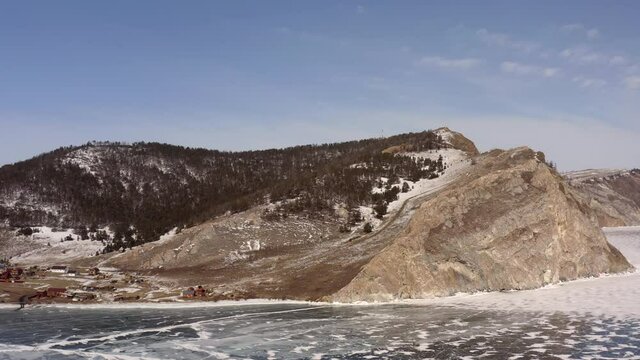 Aerial view, the drone flies forward at high speed close to the ice overlooking the village and rocky coastline. Uzury village with a coastline of rocks and hills covered with snow. Frozen Lake Baikal