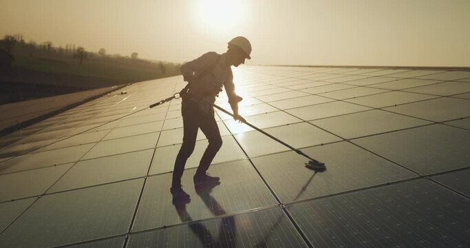 Cinematic shot of maintenance assistance technical worker in uniform is cleaning photovoltaic solar panels on roof at sunset. Concept of sustainable renewable green, bio and eco clean energy.