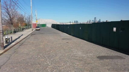 A low angle shot of a cobblestone street with a green fence. The camera dolly in, pan right and boom up to reveal an empty lot and the lower Manhattan skyline in the background on a sunny day.
