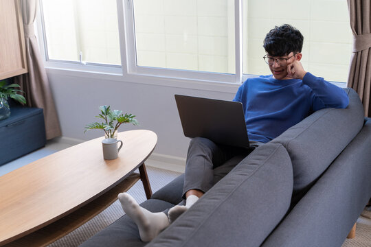 Young Asian Male Tech User Relaxing  Holding Laptop Computer And Looking At The Screen In Living Room, Remote Job Or Work From Home Concept.