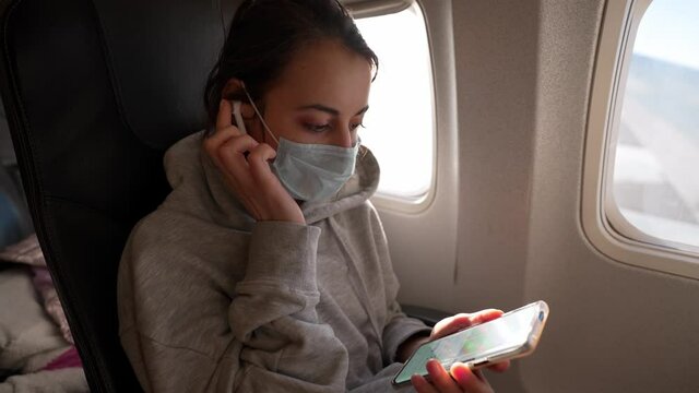 woman passenger relaxing with airpods and phone in airplane seat at window. girl in protective medical mask listen music during flight
