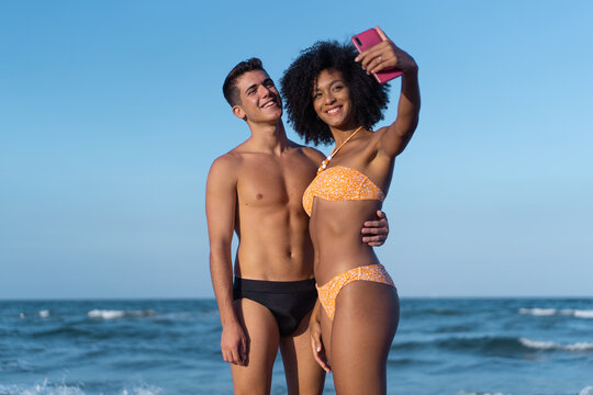 Multiracial couple in swimsuit of young people with Caucasian man and Afro-American woman taking selfie with the smartphone on the beach with the sea on the background.